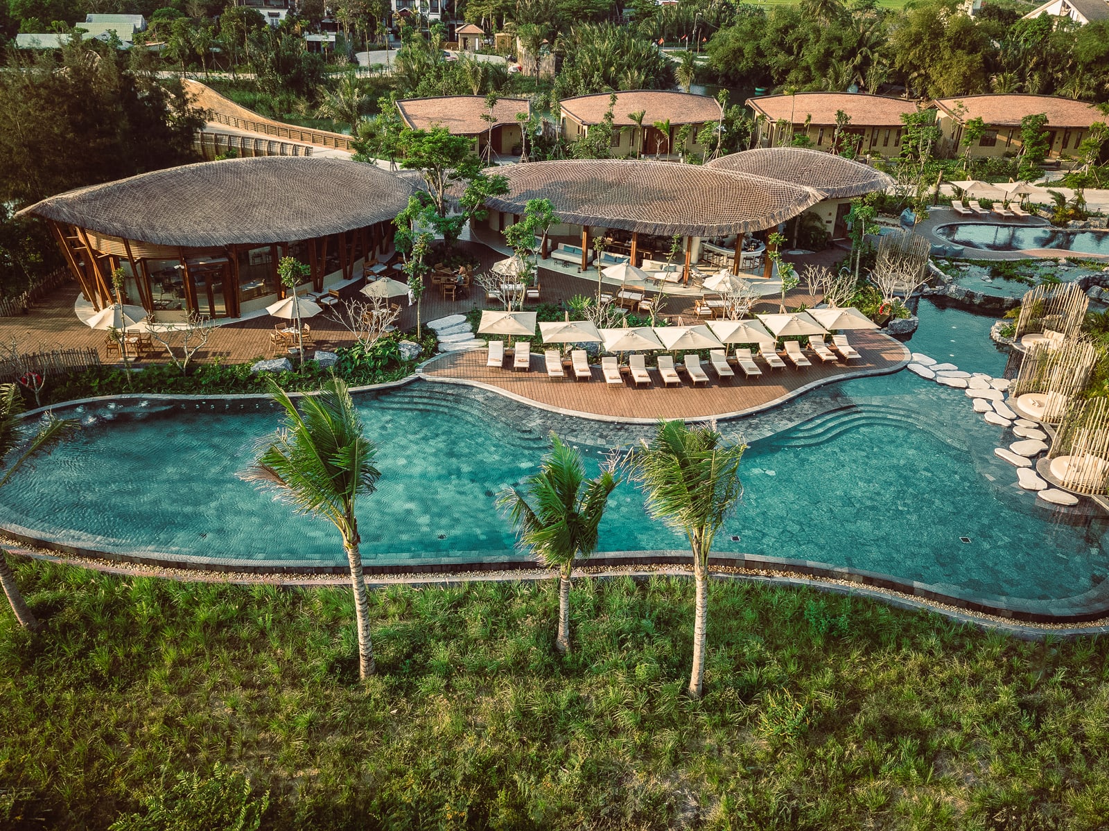 Aerial view of luxury resort with turquoise pools, thatched-roof buildings, and lounge chairs surrounded by lush greenery