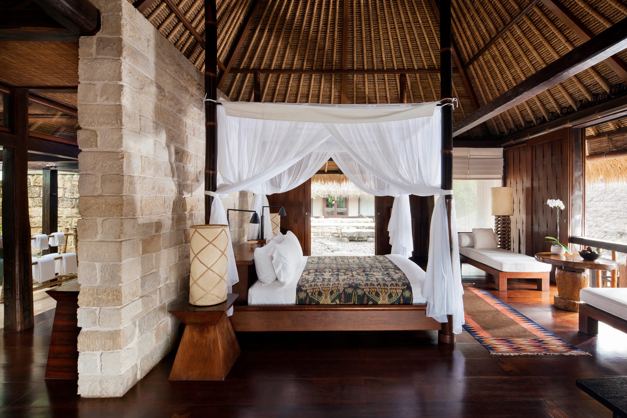 Luxury resort bedroom with four-poster canopy bed draped in white curtains, thatched ceiling, and stone column