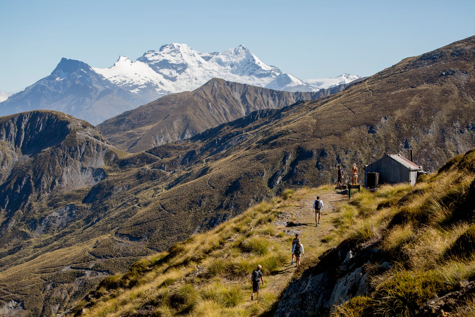 Hikers on mountain trail with snow-capped peaks and rustic shelter in distance