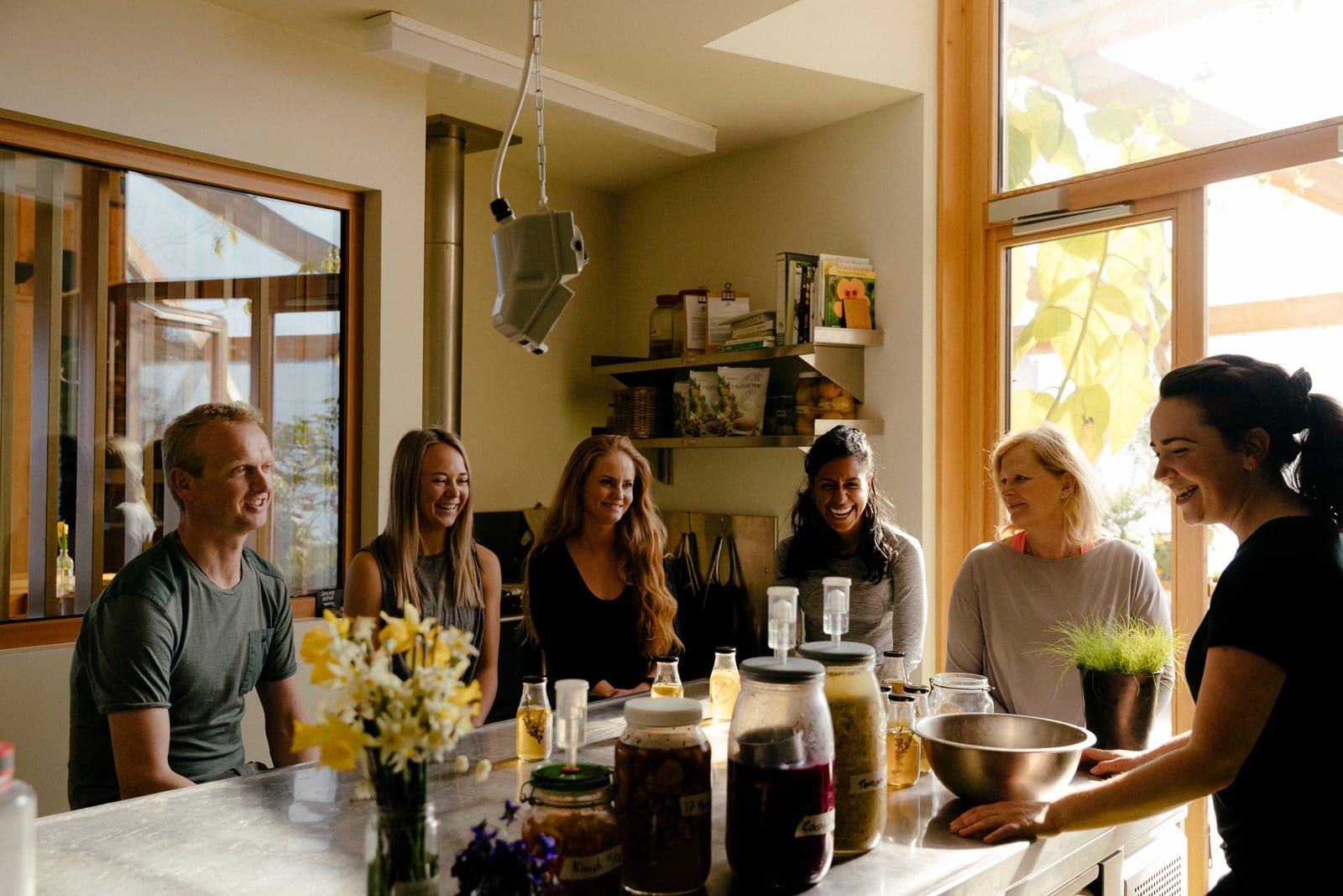 Group of six people gathered around kitchen counter with jars and flowers, natural light streaming through windows