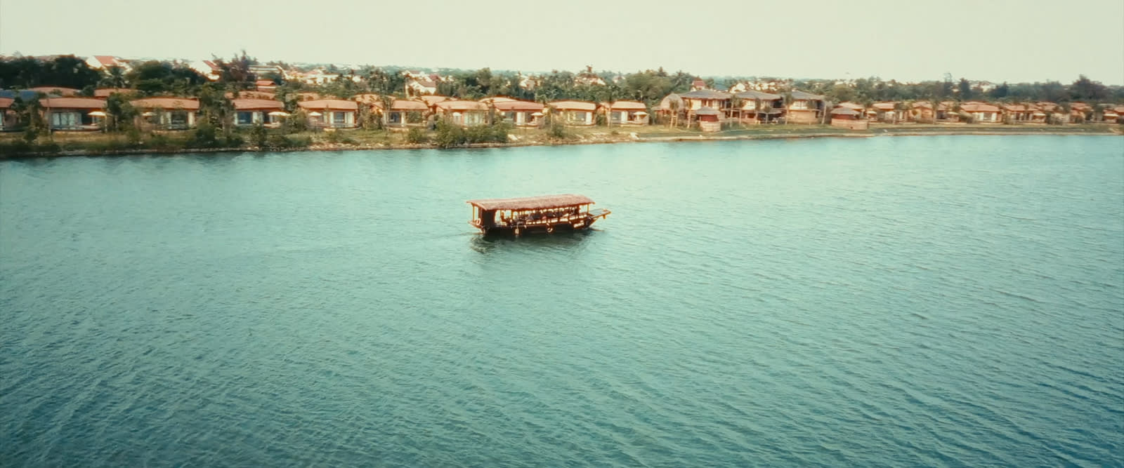Traditional wooden boat on turquoise river with waterfront villas and trees behind it