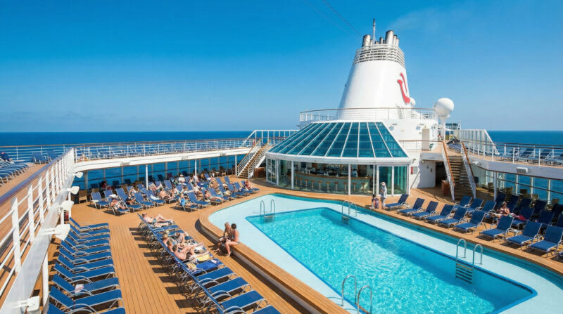 07b7ce1a-04cb-4cc1-af62-02005c9d60b8 Cruise ship pool deck with swimming pool, blue lounge chairs, and funnel in bright sunshine at sea