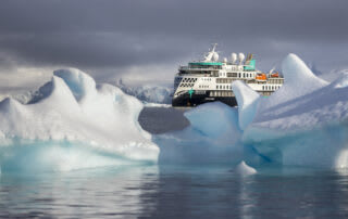 A white and teal expedition cruise ship navigating through blue-white icebergs in a misty polar