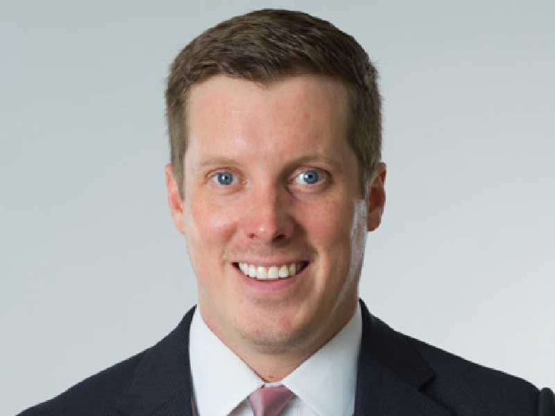 Man with blue eyes wearing dark suit and burgundy tie, smiling at camera