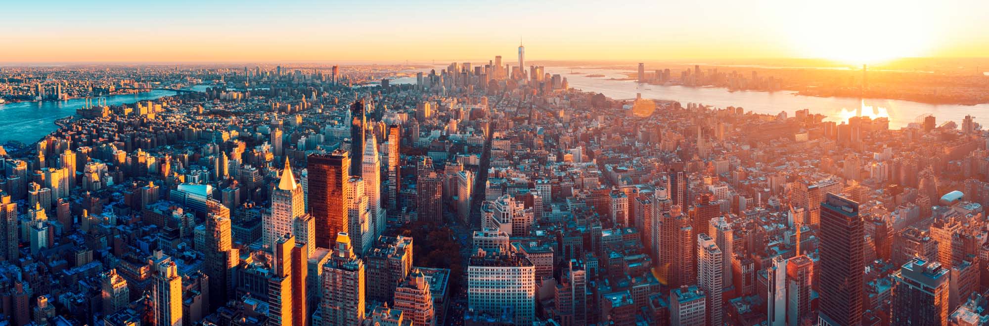 Manhattan Aerial Panorama at Sunset An expansive aerial panoramic view of Manhattan captured during golden hour, showcasing the iconic skyline with illuminated skyscrapers, the East River, Hudson River, and surrounding boroughs bathed in warm sunset light. The image captures the scale and density of New York City's architecture against a luminous golden sky.