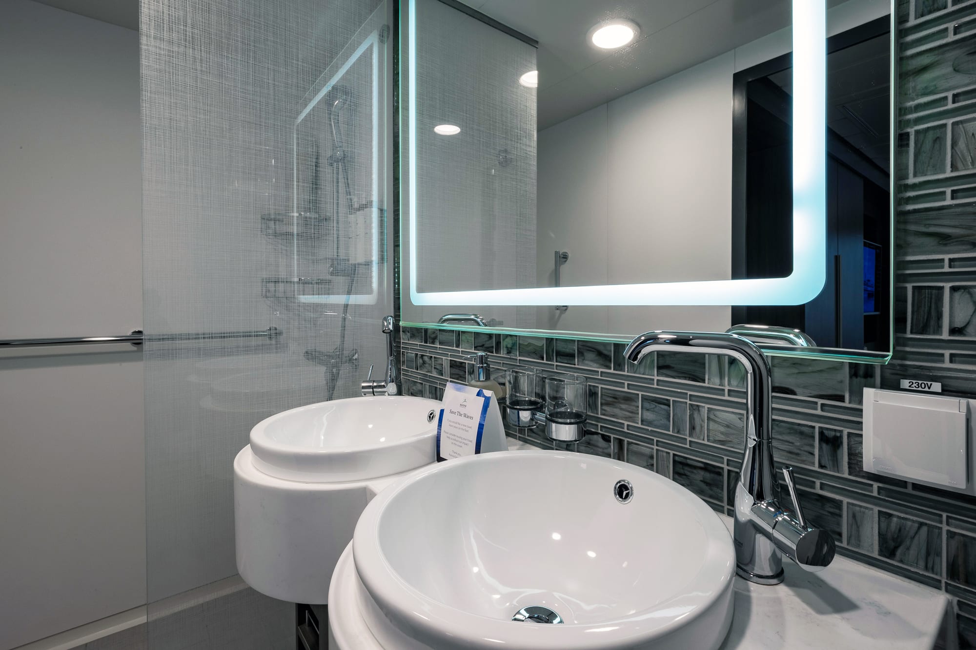 A contemporary bathroom aboard a cruise ship featuring dual white circular vessel sinks, a sleek