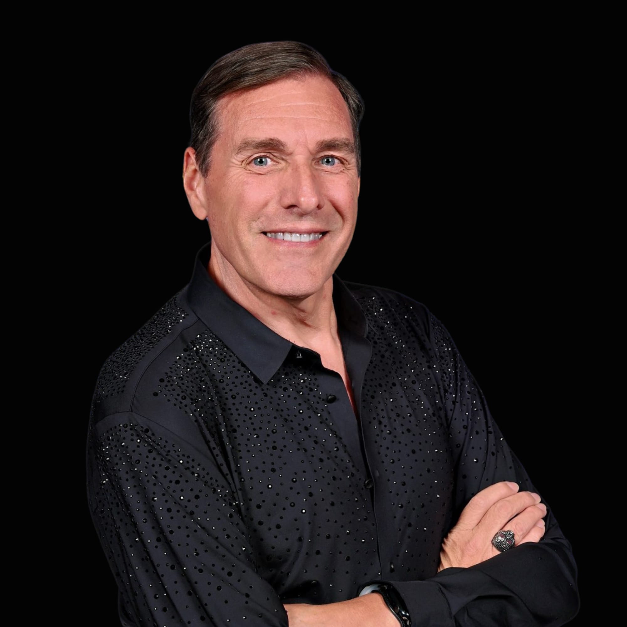 Professional Headshot Portrait A professional portrait photograph of a middle-aged man with a warm smile, wearing a black sequined/embellished button-up shirt against a black background. The image appears to be a professional headshot or promotional photo.