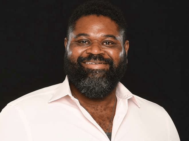 Man with full beard wearing light pink button-up shirt against dark background
