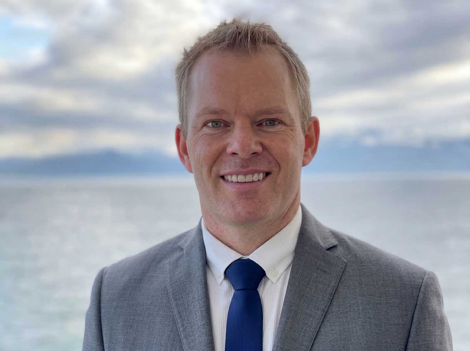 Man in gray suit and blue tie smiling at camera with ocean and cloudy sky behind him