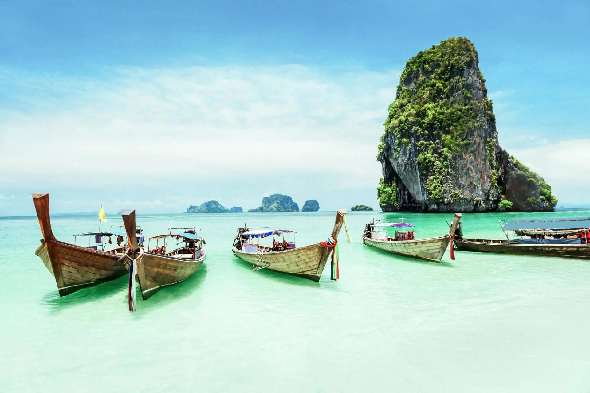 Traditional longtail boats anchored in crystal-clear turquoise water with dramatic limestone karst cliffs covered in lush vegetation rising in the background.