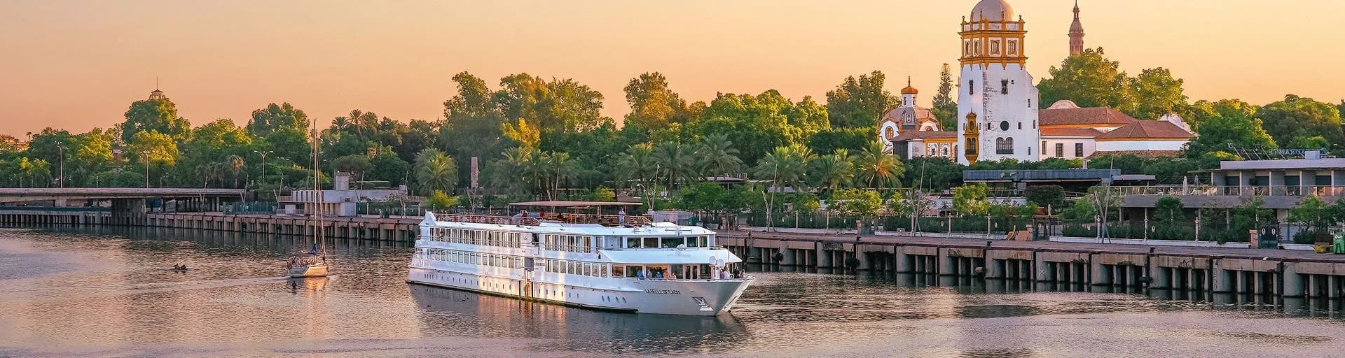 A multi-deck white riverboat moored at a wooden pier along a wide river during golden hour, with historic architecture visible on the bank.