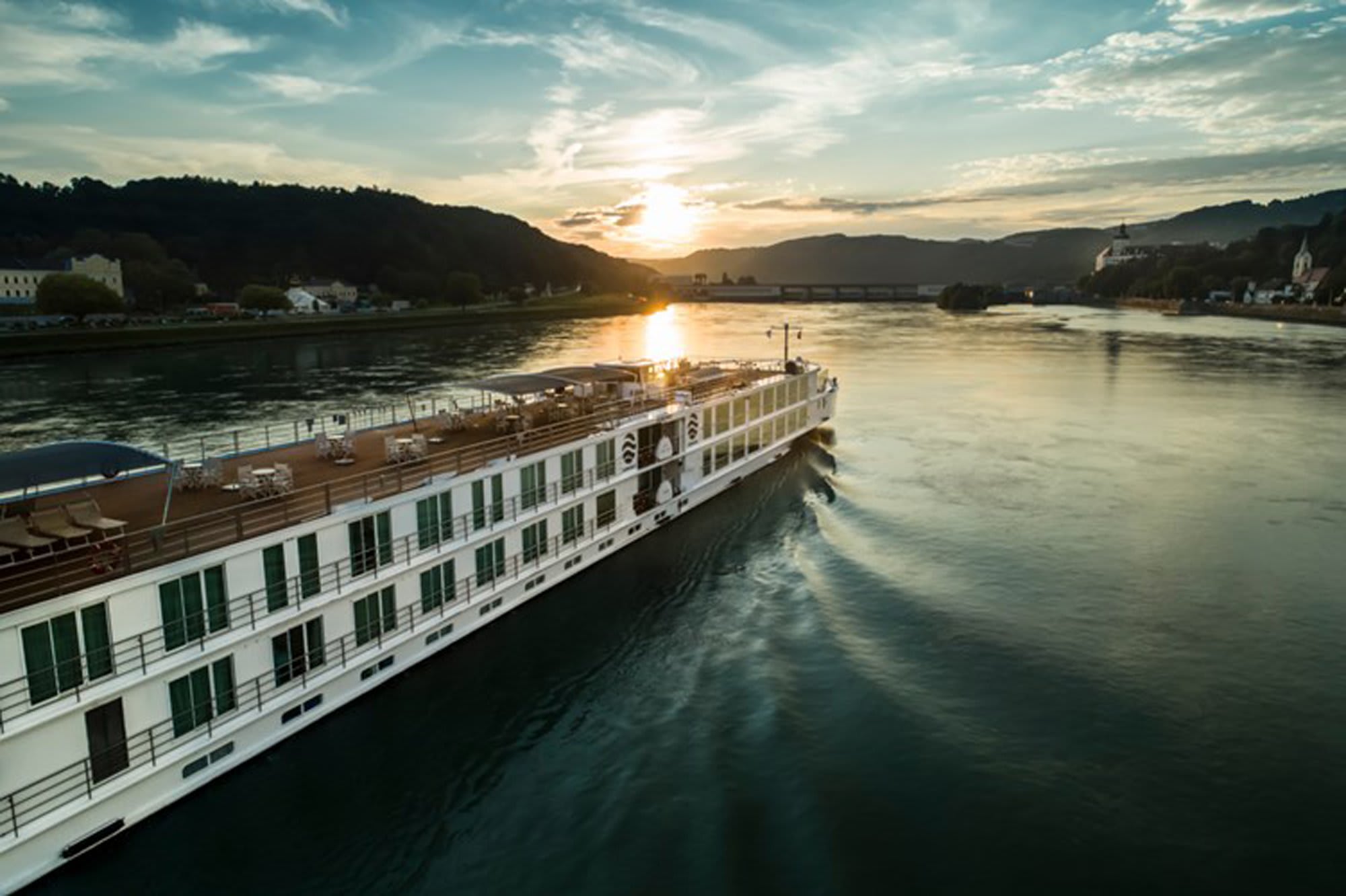 River cruise ship at sunset navigating a scenic alpine valley with forested mountains and charming riverside villages