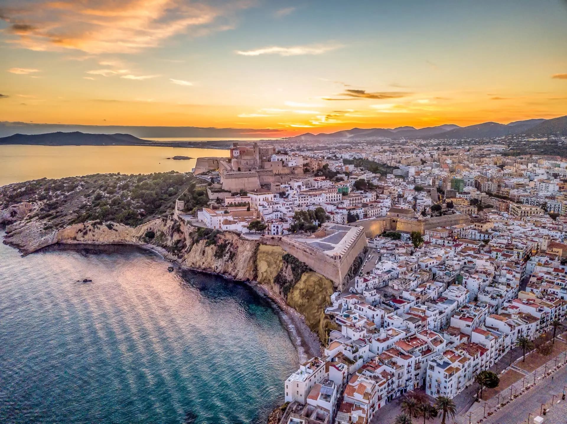 Aerial view of Denia Castle perched on a rocky peninsula overlooking the Mediterranean Sea at sunset, with white-washed buildings and mountains in the distance.