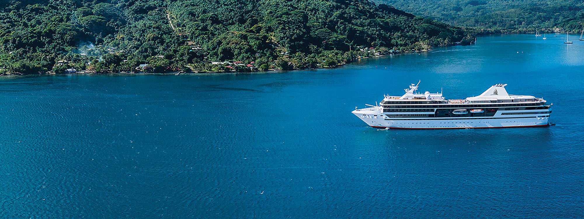 7705696 Luxury cruise ship anchored in crystalline turquoise waters alongside a lush tropical coastline in French Polynesia