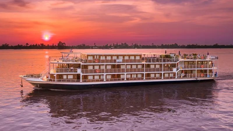 A multi-deck river cruise ship anchored on a calm river at golden hour, with a stunning sunset painting the sky in shades of orange, pink, and purple.