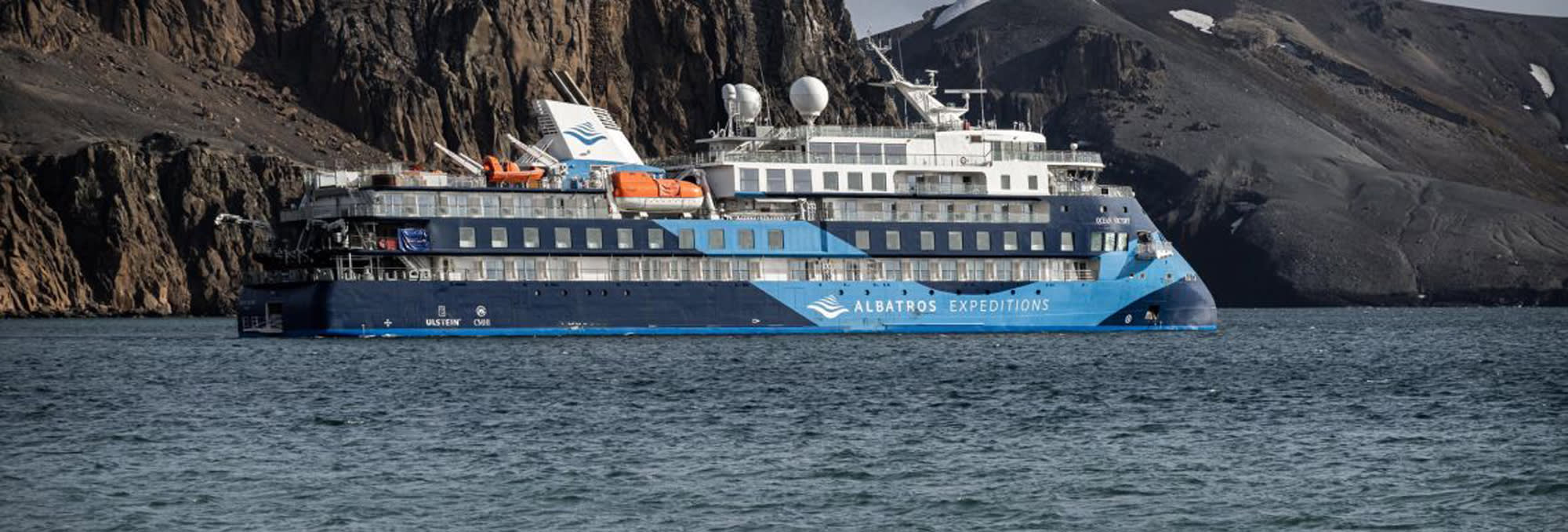 An expedition cruise ship from Polar Latitudes anchored near a rugged, rocky coastline with steep mountainous terrain in the background.