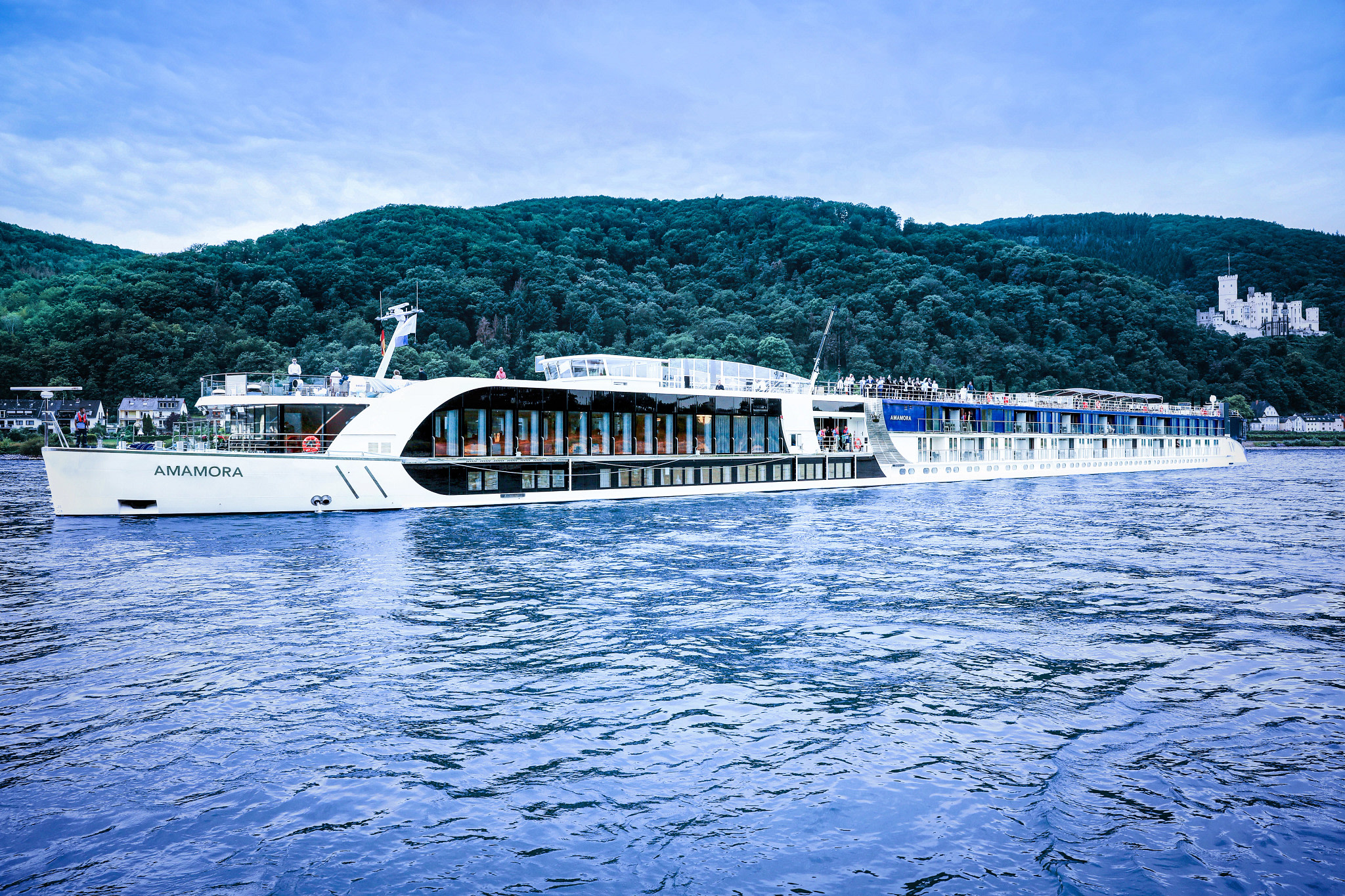 AmaMora River Cruise Ship on Scenic Waterway A modern river cruise ship named AmaMora sailing on a blue river with lush green forested hills and a white castle in the background. The vessel features large panoramic windows and a sleek white design.