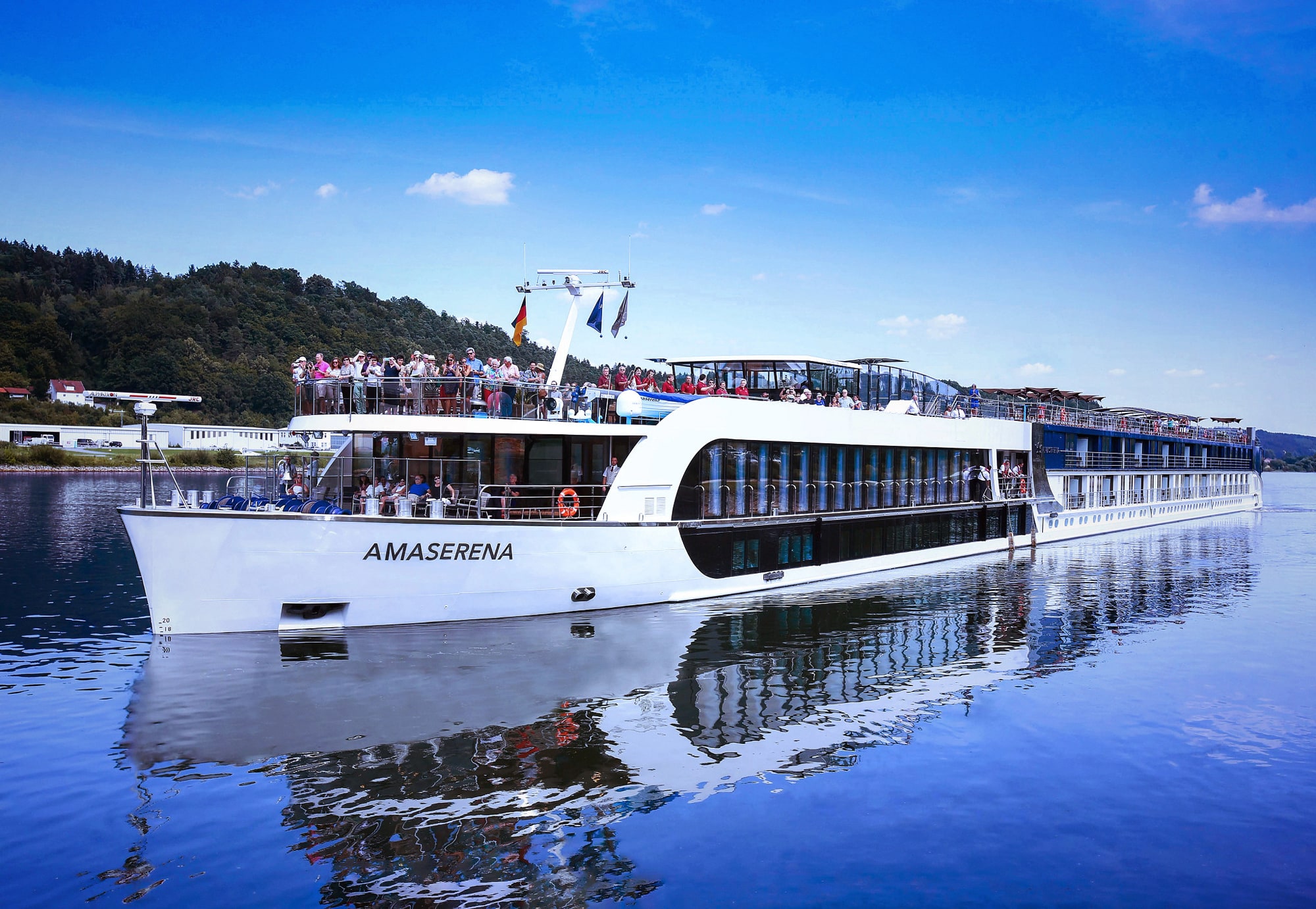AmaSerena River Cruise Vessel on Calm Waters A modern white river cruise ship named AmaSerena docked on a serene blue river, with passengers visible on multiple decks against a backdrop of forested shoreline and blue sky.