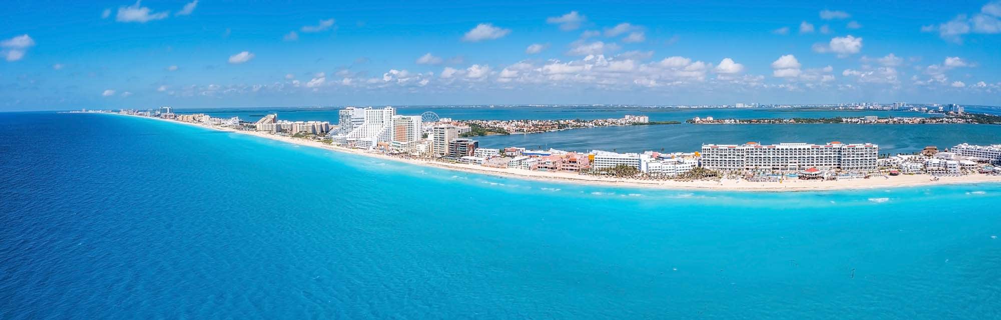 A panoramic aerial view of Cancun's stunning coastline, featuring turquoise waters, white sandy beaches, and a dense line of high-rise hotels and resorts along the shoreline.