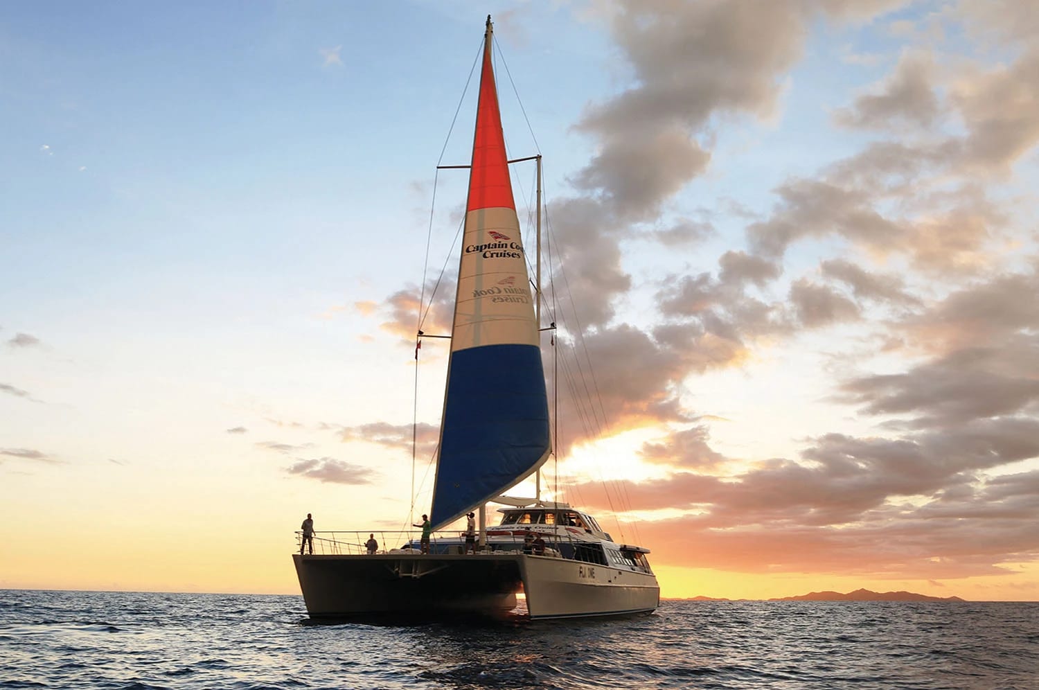 Captain Cook’s Catamaran Sunset Cruise A Captain Cook Cruises catamaran with distinctive red, tan, and blue sails glides across calm ocean