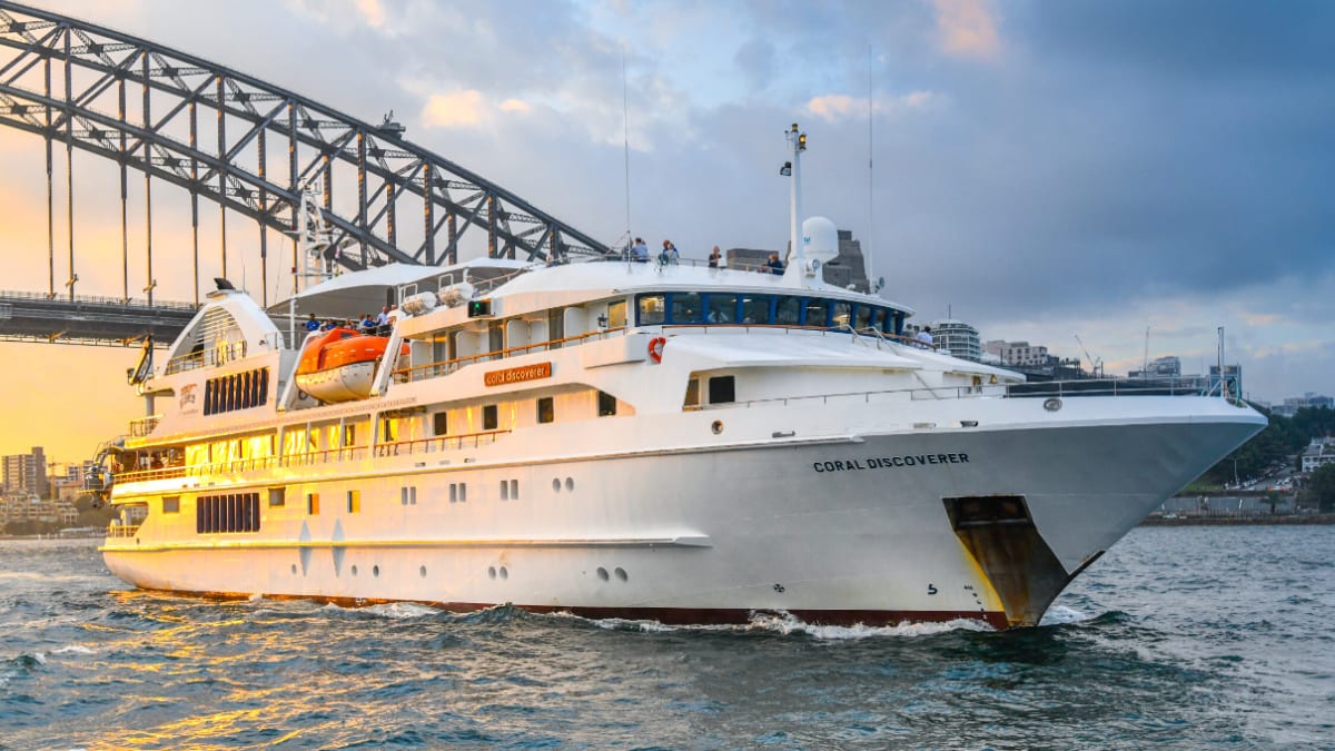 A white expedition cruise ship named Coral Discoverer navigating through Sydney Harbour with the iconic Harbour Bridge in the background during golden hour lighting.