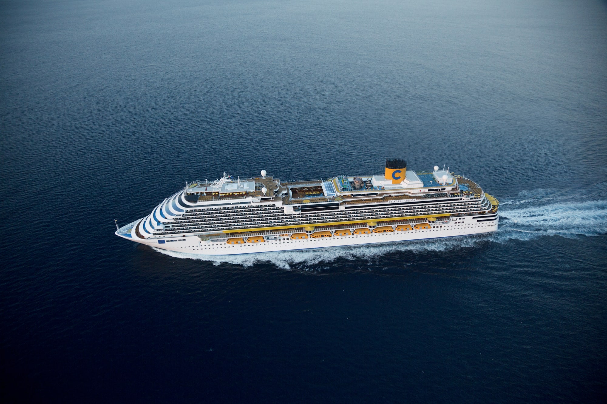 A large white and yellow Costa cruise ship sailing across a calm, deep blue ocean, showcasing the vessel's sleek design and impressive size.