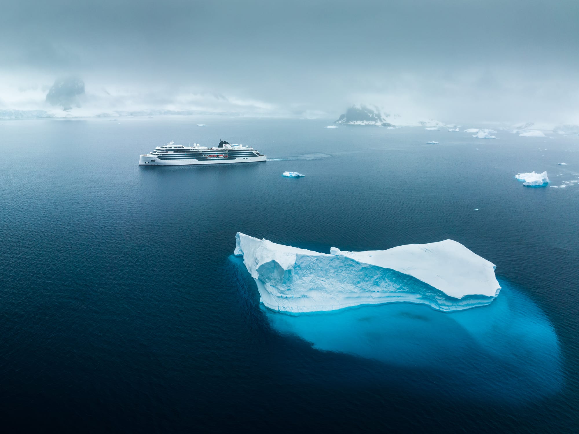 Cruise Ship Amidst Polar Icebergs A modern white cruise ship sailing through cold arctic waters near large, blue-white icebergs with misty mountainous islands in the background.