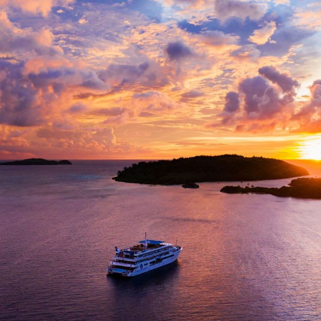 A modern cruise ship is anchored in calm turquoise waters during a spectacular golden and purple sunset, with a lush forested island silhouetted in the background and dramatic cloud formations illuminated by the setting sun.