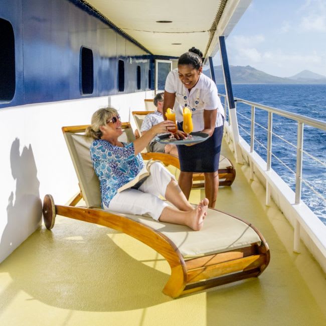 A crew member serves refreshments to a passenger relaxing on a lounge chair on the promenade deck
