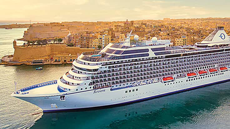 Cruise Ship Docked in Mediterranean Port A large white cruise ship anchored near a historic coastal city with golden stone buildings and fortifications. The vessel is positioned in turquoise waters during golden hour lighting.