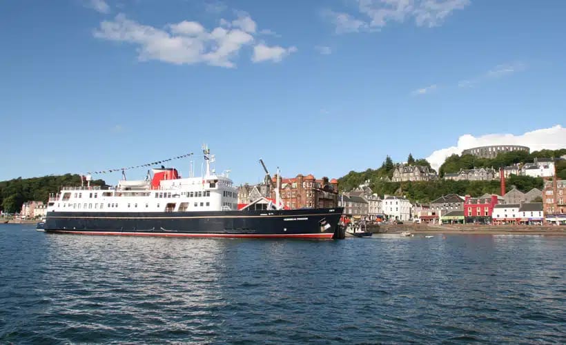 A large passenger cruise ship with distinctive red and white livery is moored in the picturesque Scottish port town of Oban, with colorful waterfront buildings and a hilltop structure visible in the background under clear blue skies.
