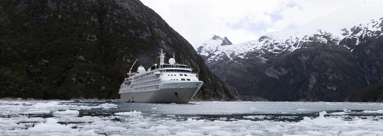 A white cruise ship sailing through ice-filled waters surrounded by dramatic snow-capped mountains