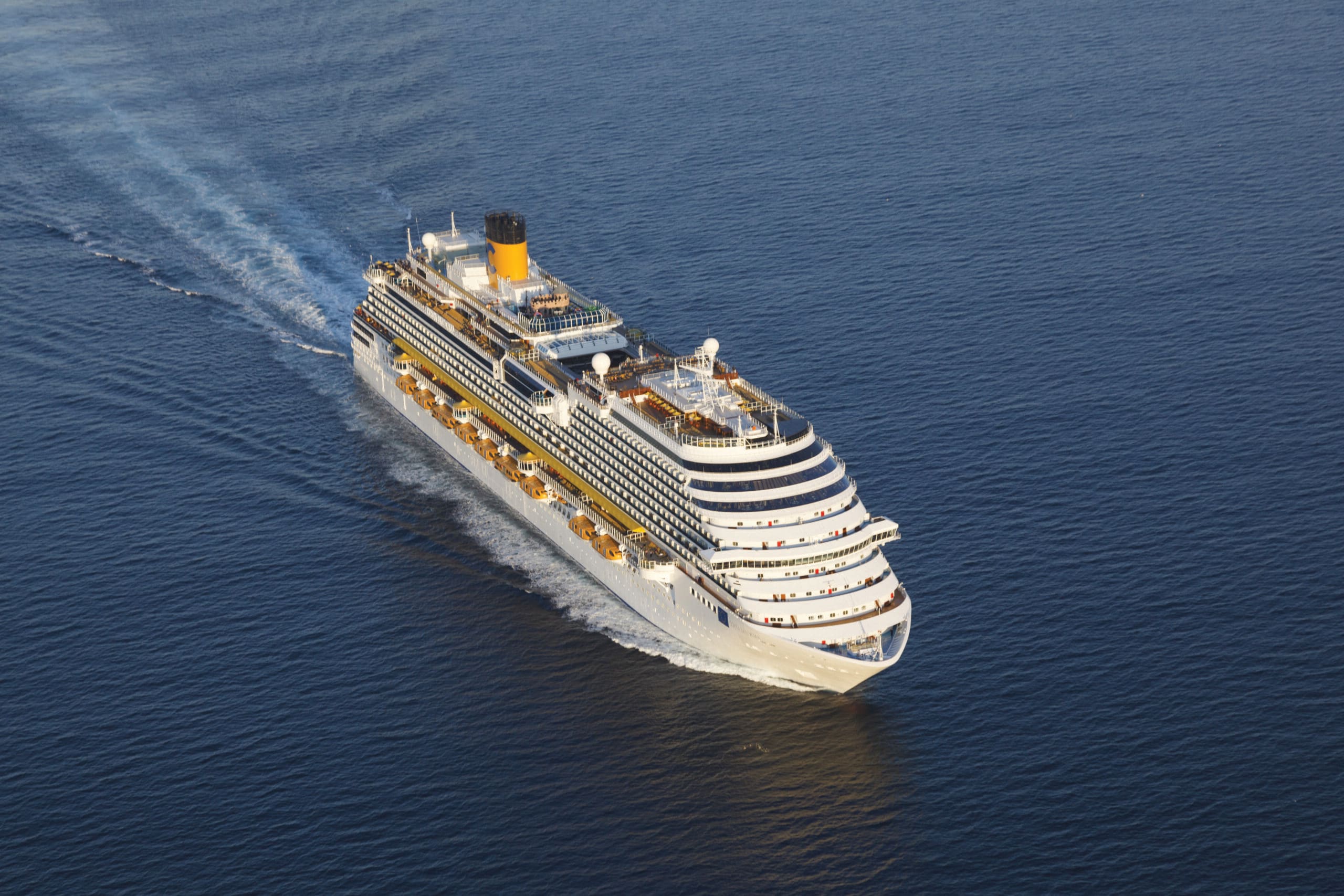 A large white and yellow cruise ship sailing across a deep blue ocean, creating a distinctive wake