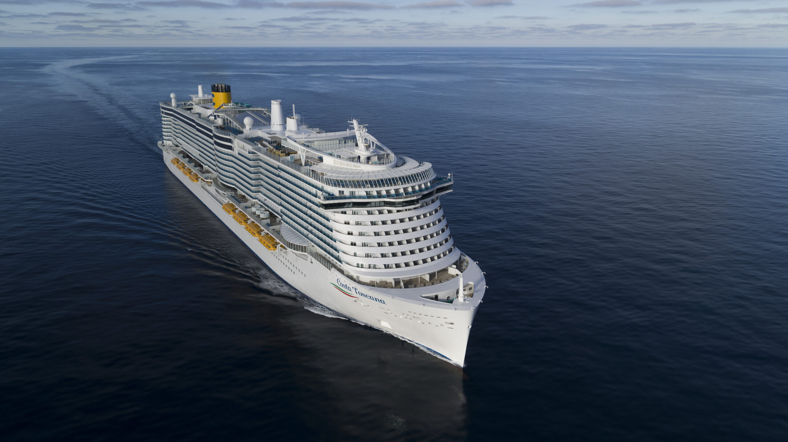 A large white cruise ship with yellow and white color scheme sailing across a serene, flat blue ocean under an overcast sky.
