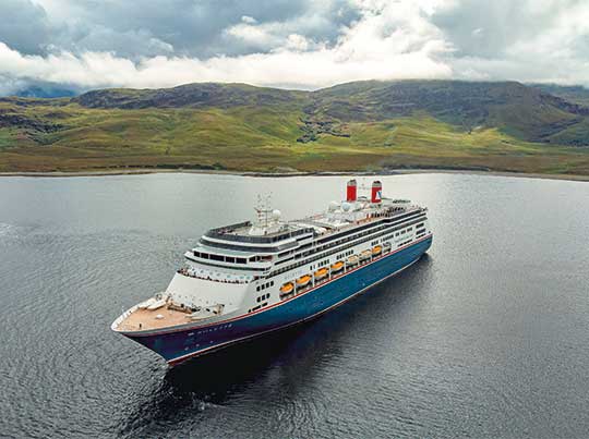 A large modern cruise ship with distinctive red and blue hull markings anchors in calm waters off