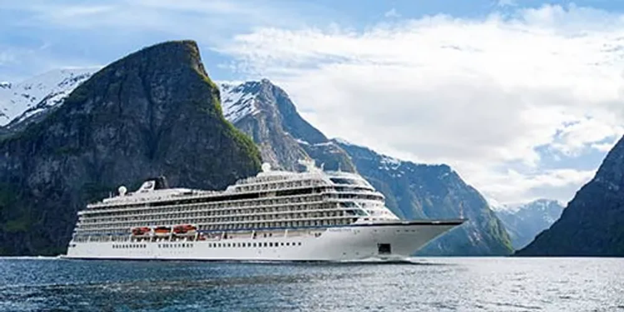 A large white cruise ship sailing through a stunning fjord with steep, snow-capped mountains