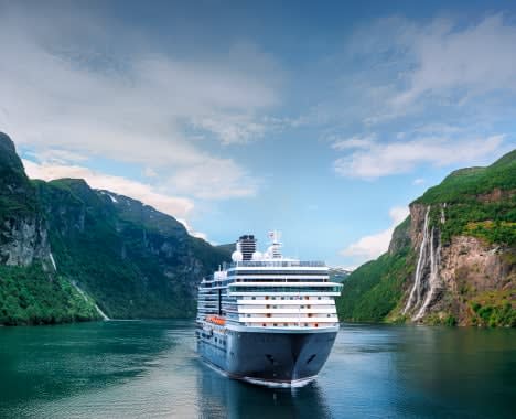 A white cruise ship sailing through a dramatic fjord landscape with steep, green-covered mountains and multiple waterfalls cascading down rocky cliffs.