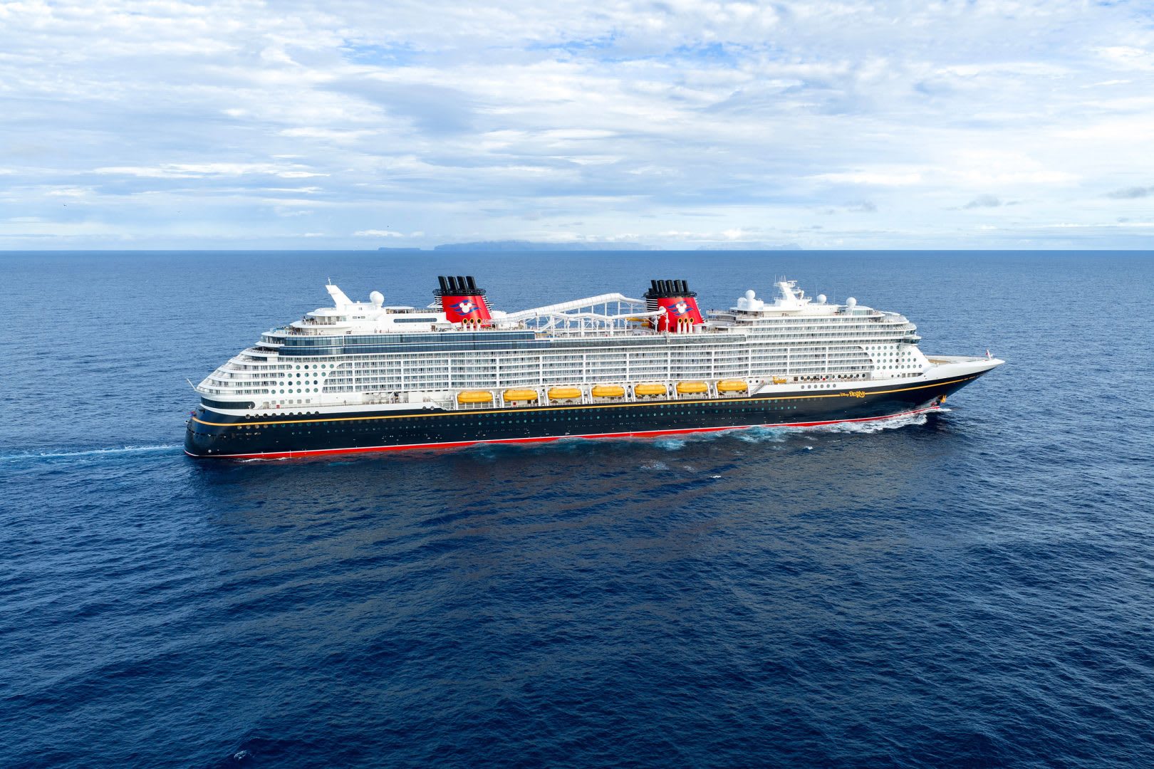 Disney Cruise Line Ship at Sea A massive contemporary cruise ship navigates open ocean waters under a partly cloudy sky with distinctive red funnels and dark blue hull.