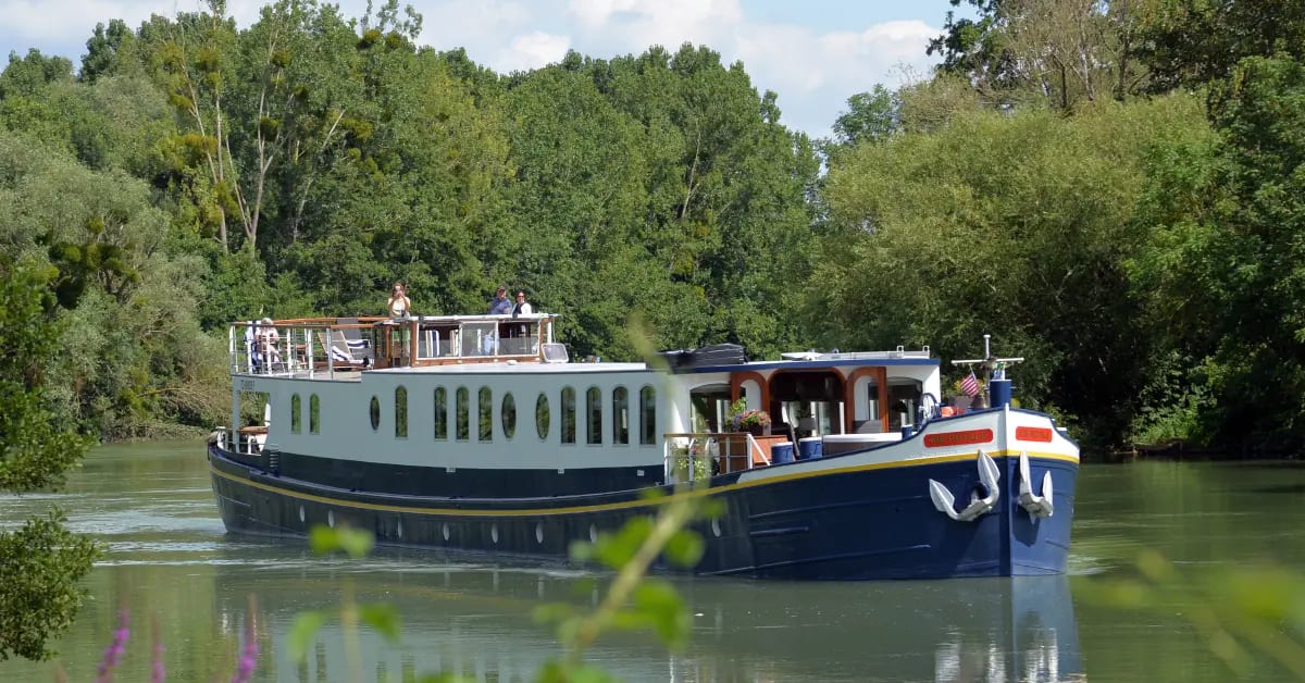 European River Cruise Boat on Waterway A charming European river cruise barge with distinctive blue and white hull navigates a calm waterway surrounded by lush green trees and vegetation.