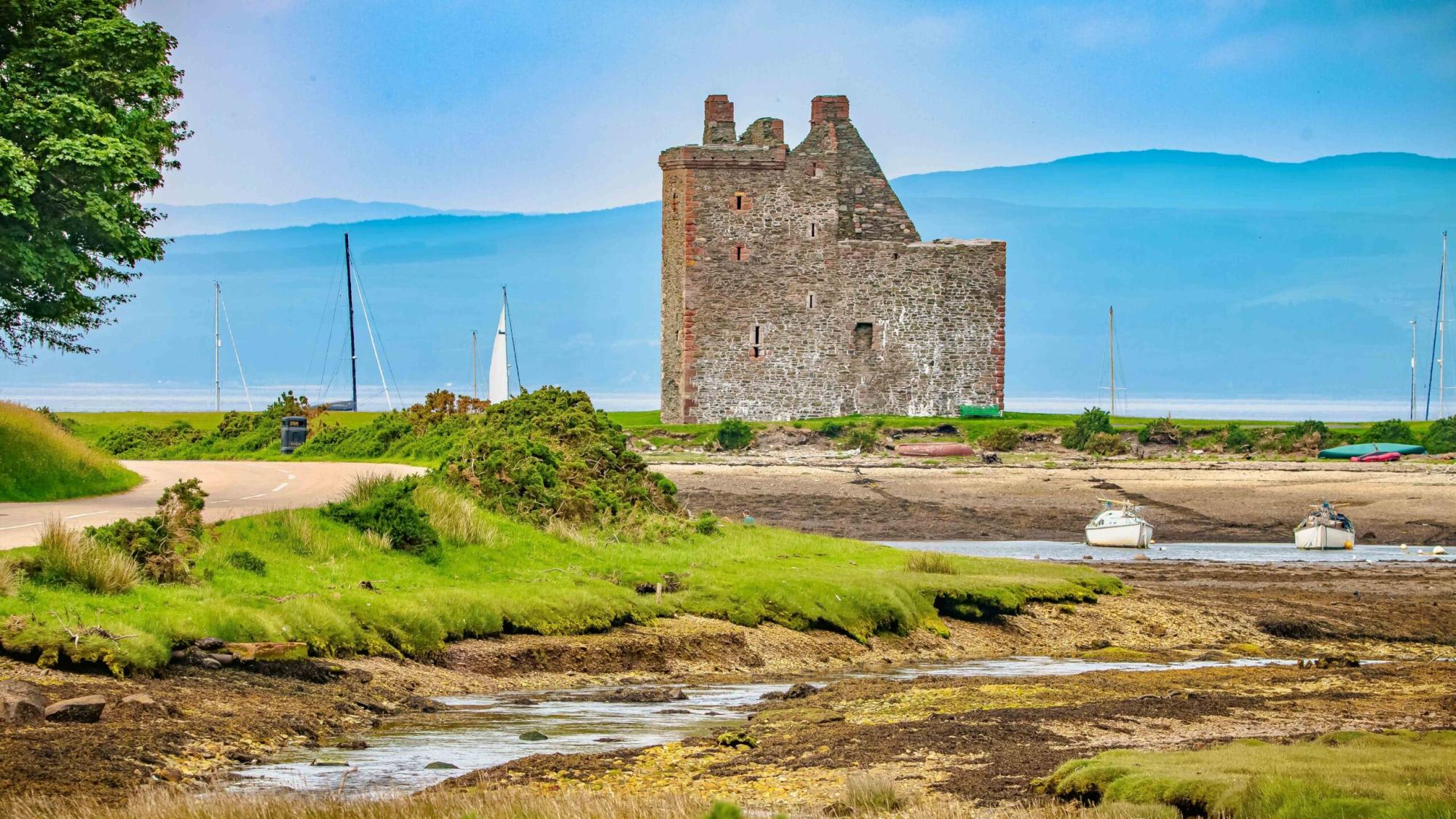 A picturesque medieval stone castle tower stands on the banks of a Scottish estuary, surrounded