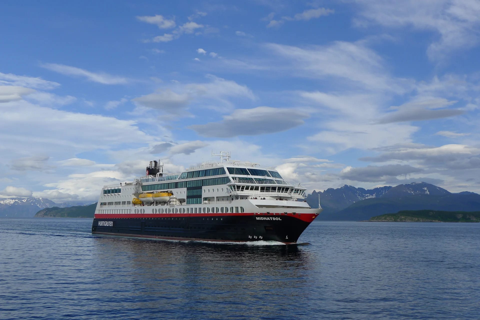 Hurtigruten Cruise Ship in Norwegian Fjords A Hurtigruten cruise ship navigating through a scenic Norwegian fjord landscape with snow-capped mountains in the background under a dramatic cloudy sky.