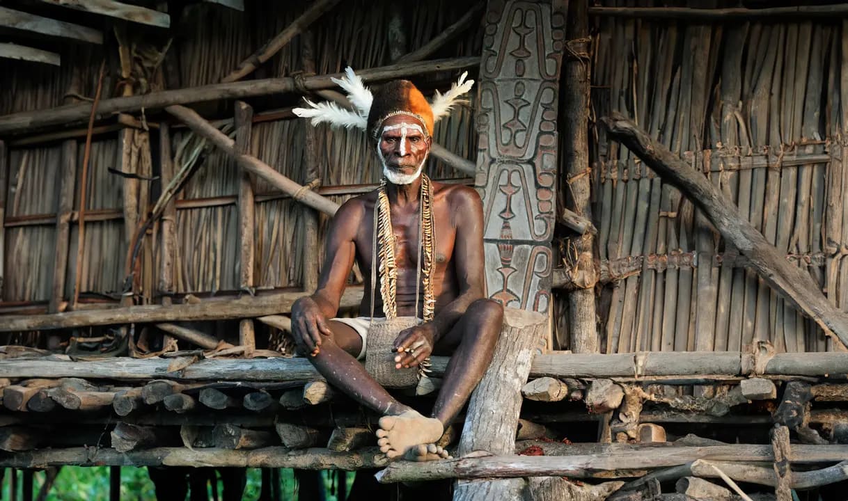 A dignified tribal elder from Papua New Guinea sits on a traditional wooden structure, displaying