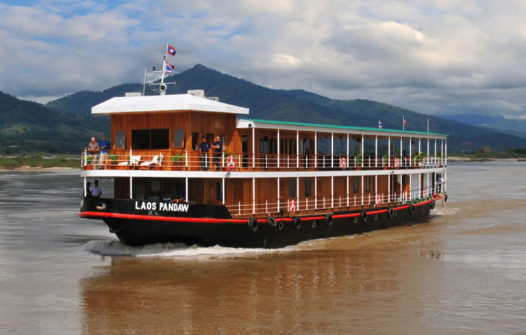 A wooden river cruise boat named Laos Pandaw navigating through a scenic river with mountainous landscape in the background. The multi-deck vessel appears to be traveling on the Mekong River in Southeast Asia.