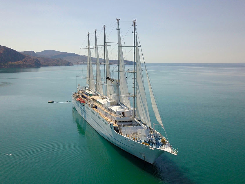 A multi-masted sailing vessel anchored in tranquil blue-green waters with mountainous coastline in the background.