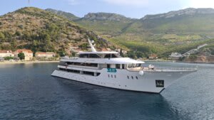 A modern expedition-style cruise ship is anchored in calm turquoise waters near a picturesque