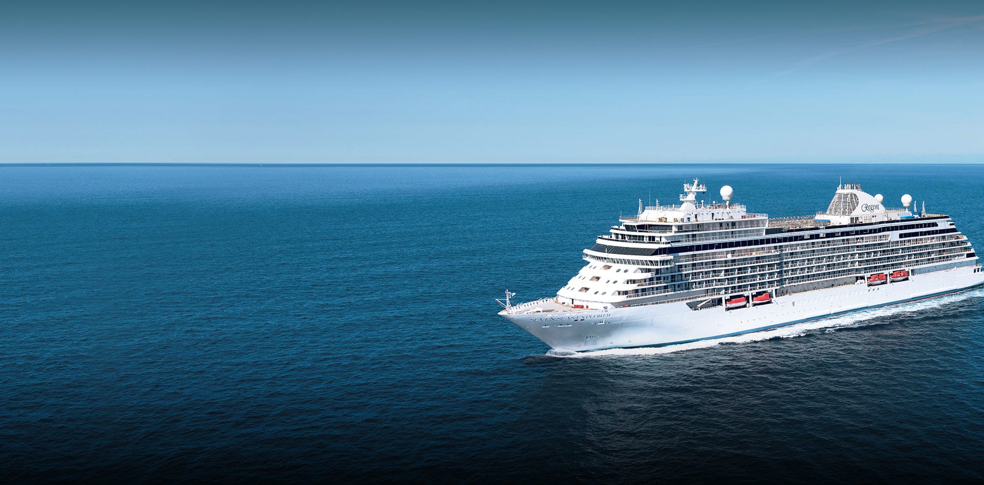 A pristine white luxury cruise ship sailing across a tranquil, deep blue ocean under a clear blue sky. The vessel appears to be moving at speed, creating a white wake against the calm sea.