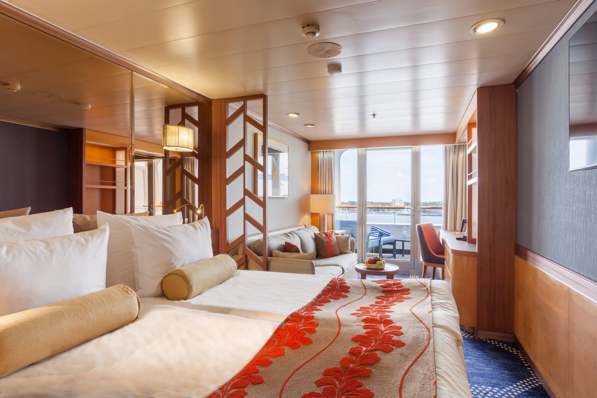 An elegant cruise ship cabin featuring a spacious bedroom with premium furnishings, wooden accents, and a separate living area with ocean view balcony overlooking the sea. The suite exemplifies modern cruise ship luxury accommodations with warm earth tones and contemporary design elements.