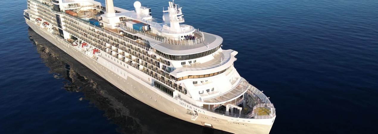 A large white cruise ship sailing on calm blue waters, showcasing multiple decks and modern design. The vessel appears to be a high-end luxury liner with gleaming white exterior and multiple levels of balconies.