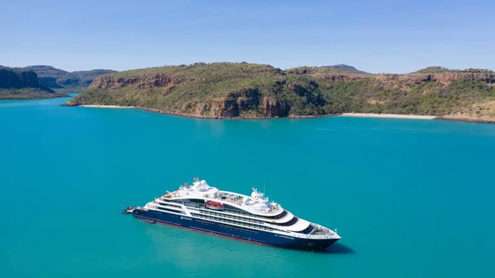 A sleek, modern cruise ship anchored in stunning turquoise waters near a rugged, green coastal
