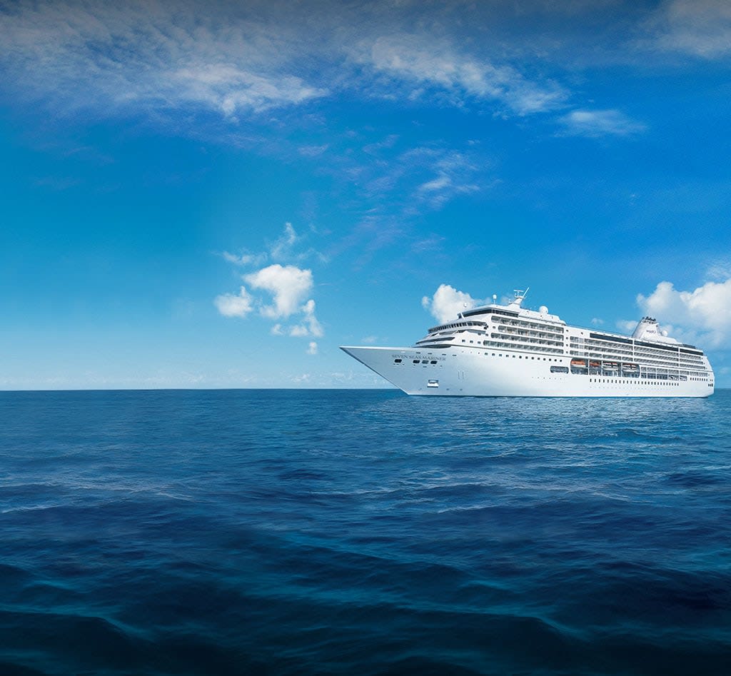 A pristine white cruise ship sailing on deep blue waters under a clear sky with scattered white clouds. The vessel appears large and elegant, representing modern maritime travel.